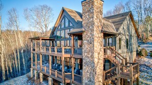 A cabin with a stone chimney, multiple decks, and a view of the surrounding forest.
