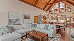 A spacious living room with a large sectional sofa, a wooden coffee table, and a rug. There is a kitchen area with wooden cabinets and a dining table. The room has a high ceiling with exposed wooden beams and large windows.