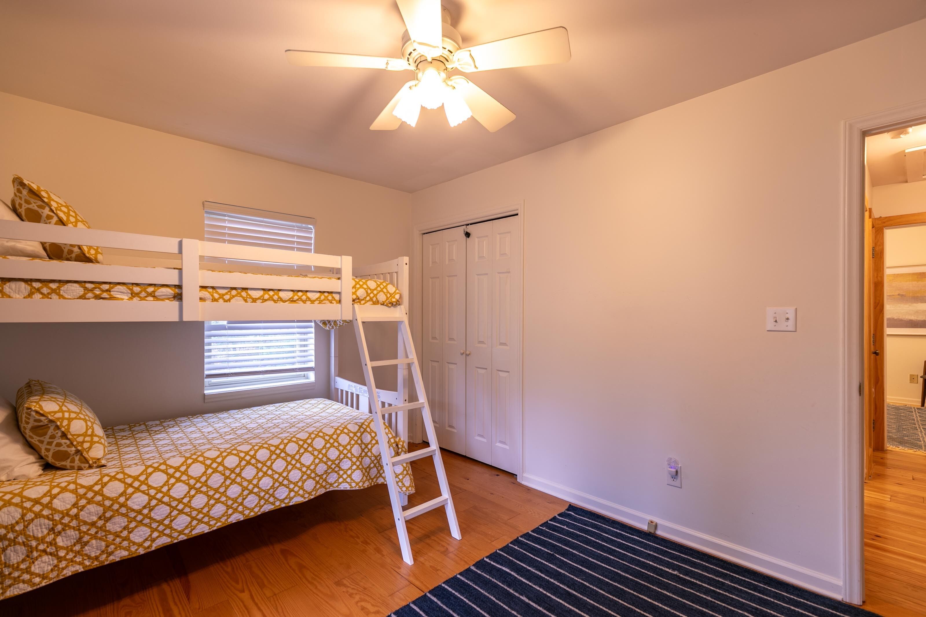 A room with bunk beds, a ceiling fan, a window with blinds, a door, and a wooden floor.