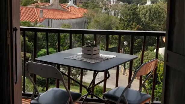 Un balcon avec une table et des chaises, donnant sur un paysage côtier avec des bâtiments et des arbres.