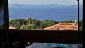 Une vue depuis une fenêtre donnant sur une zone côtière, avec des voiliers sur la mer, des arbres verdoyants et des toits en tuiles rouges.