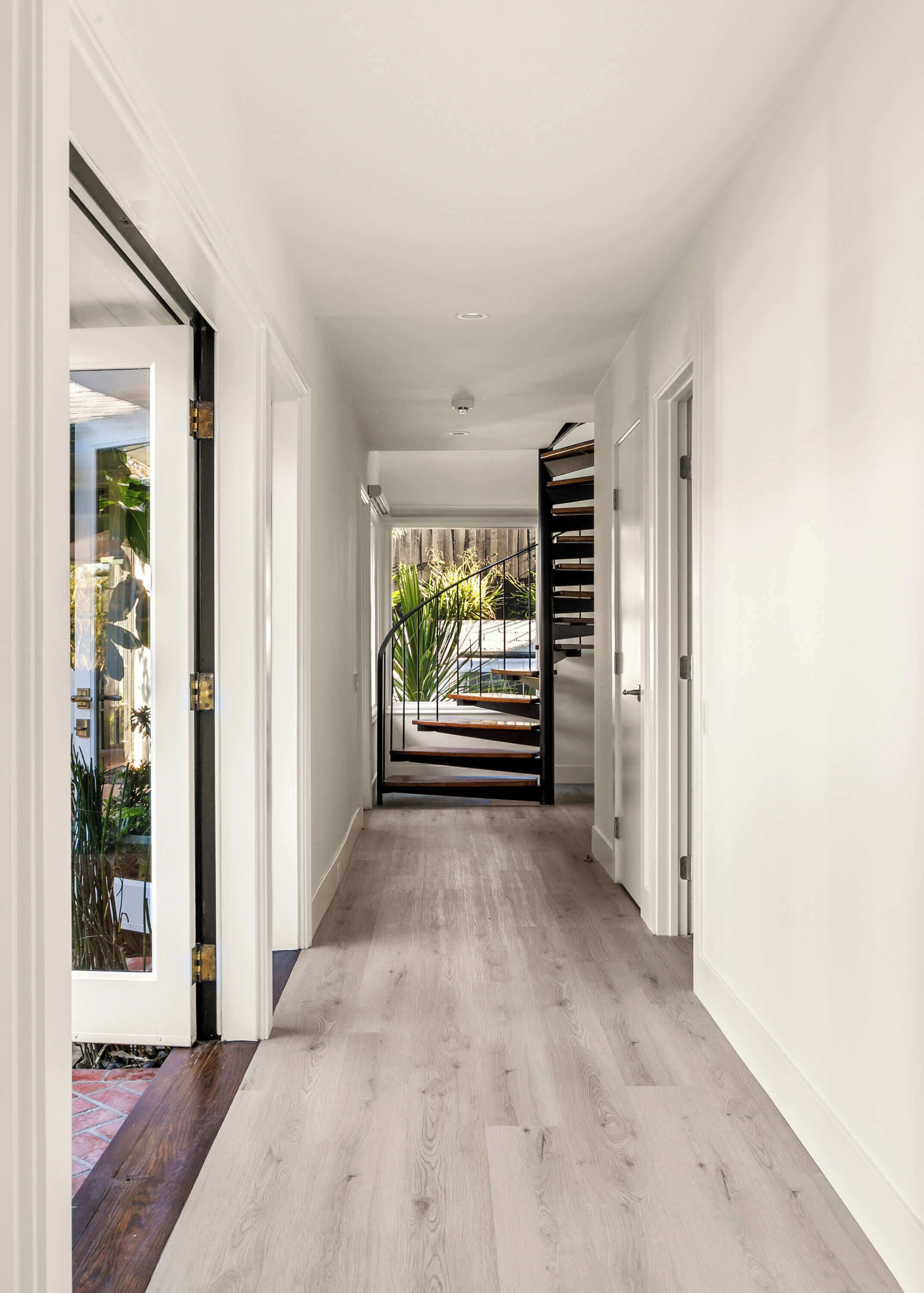 A hallway with a wooden floor, white walls, and a staircase leading to an outdoor area with plants.