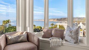 A room with a large bay window overlooking a coastal view, featuring a cushioned seating area with pillows and a throw blanket.