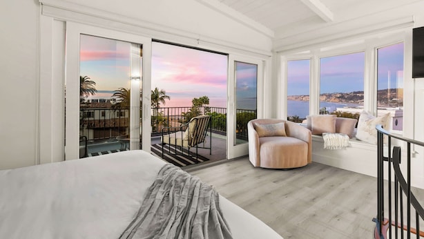 A bedroom with a large bed, a balcony with a view of the sea, and a seating area with a chair and cushions.