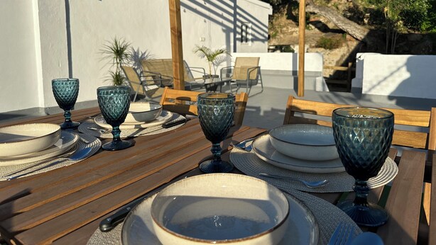 A table set outdoors with plates, bowls, and glasses, surrounded by wooden chairs and a pergola. - Casa rural Los Alcornocales (Alcala de los Gazules)