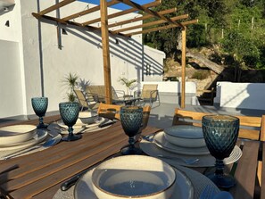 A table set outdoors with plates, bowls, and glasses, surrounded by wooden chairs and a pergola. - Casa rural Los Alcornocales (Alcala de los Gazules)