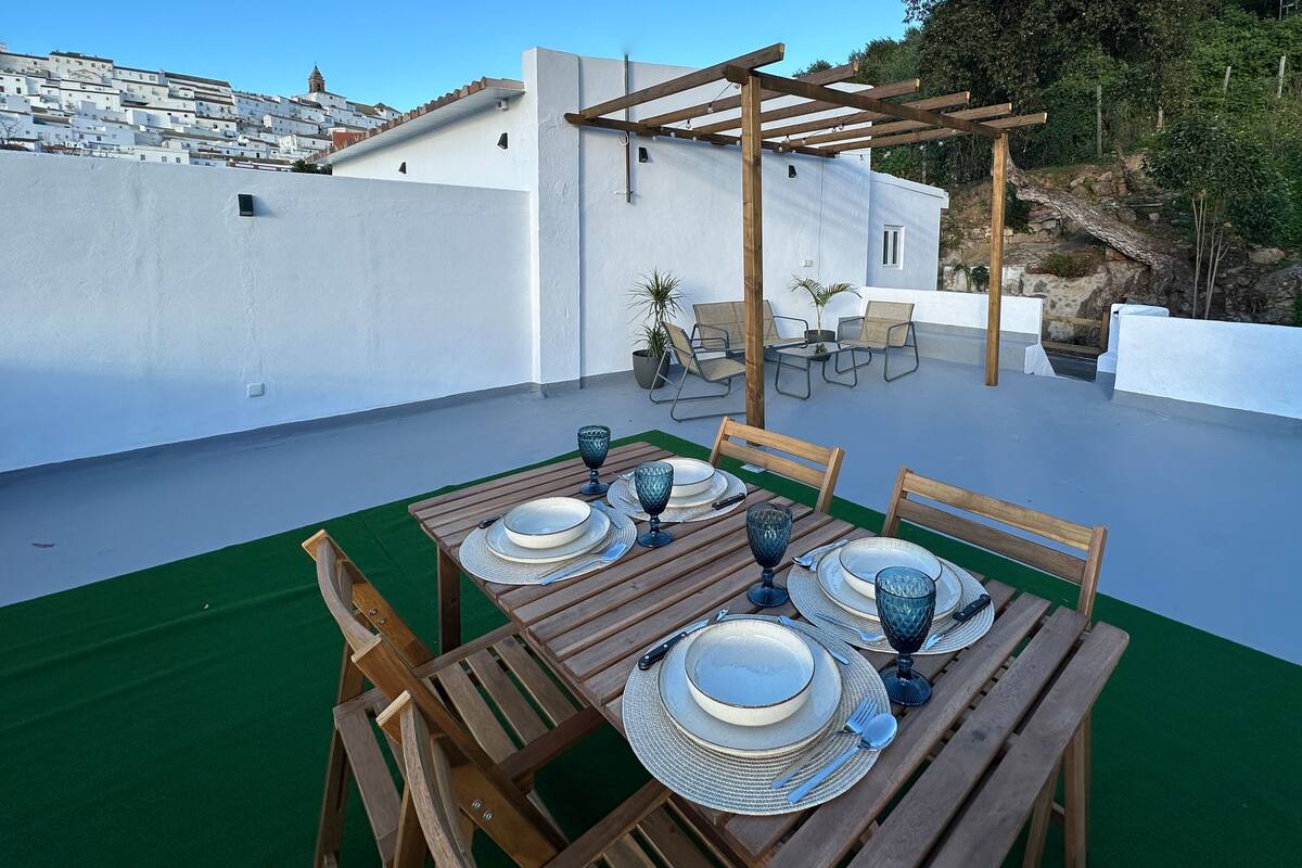 A rooftop terrace with a wooden table set for four, surrounded by chairs, and a pergola structure.