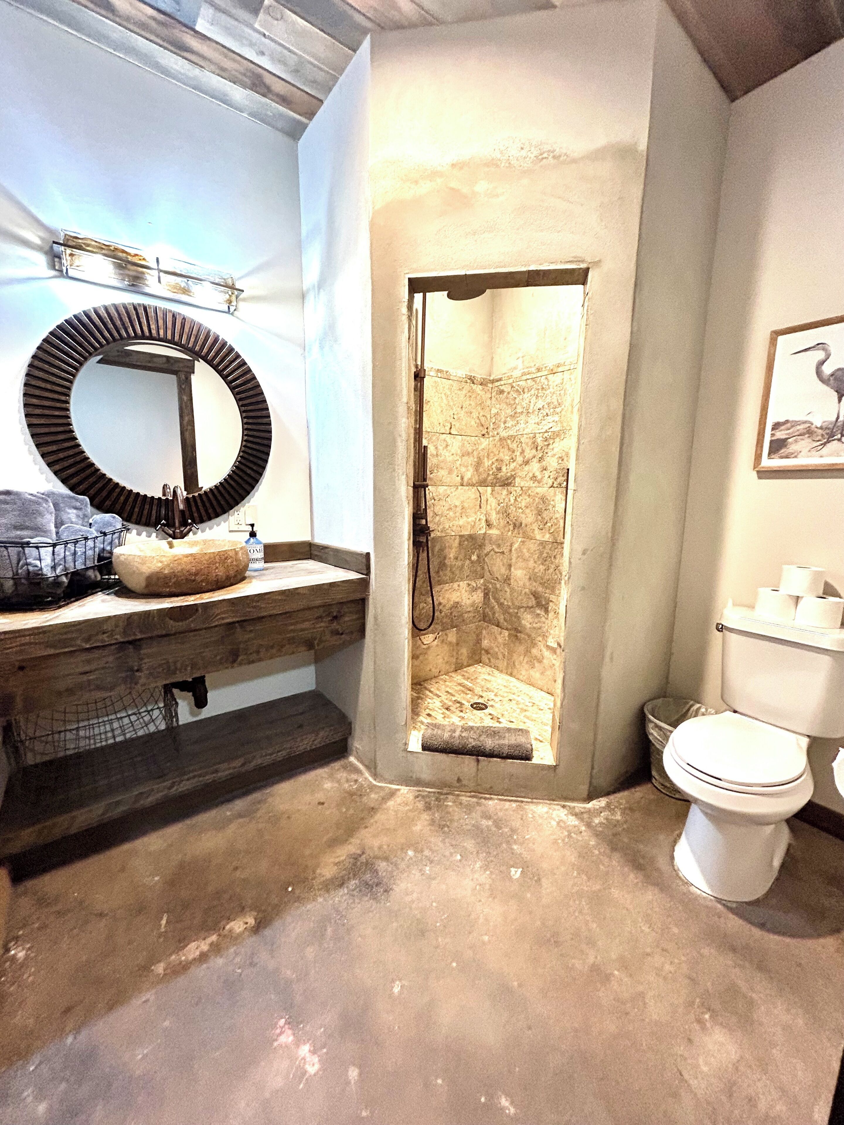 A bathroom with a wooden vanity, a round mirror, a shower with a stone wall, and a toilet.