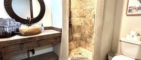 A bathroom with a wooden vanity, a round mirror, a shower with a stone wall, and a toilet.