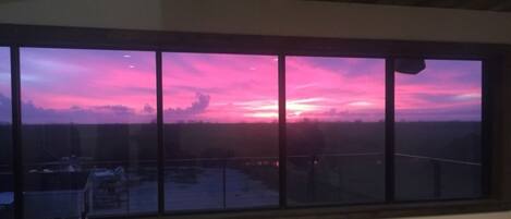 Een groot raam met uitzicht op een zonsondergang boven een vlakke omgeving.