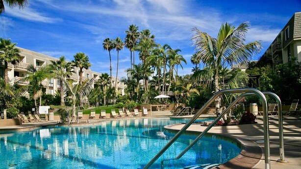 Une piscine entourée de chaises longues et de palmiers, le tout adossé à un bâtiment d’hôtel.