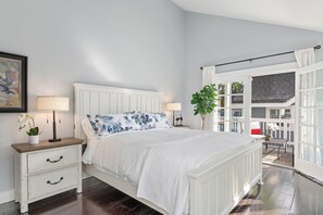 Une chambre avec un lit blanc, deux tables de chevet avec des lampes, une fenêtre avec des rideaux et une vue sur l’extérieur.