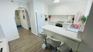 Une cuisine moderne avec des meubles blancs, un réfrigérateur en acier inoxydable, un four à micro-ondes et un évier. On y trouve également un coin bar avec des tabourets et une petite plante posée sur le comptoir.