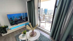 Un salon moderne doté d’un grand téléviseur à écran plat, d’une petite table ronde ornée d’un vase et de plantes, et d’un balcon offrant une vue sur la ville.