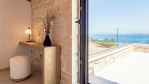 Un muro de piedra, un banco de madera, un taburete blanco y un puerta de cristal que da a un balcón con vista al mar.