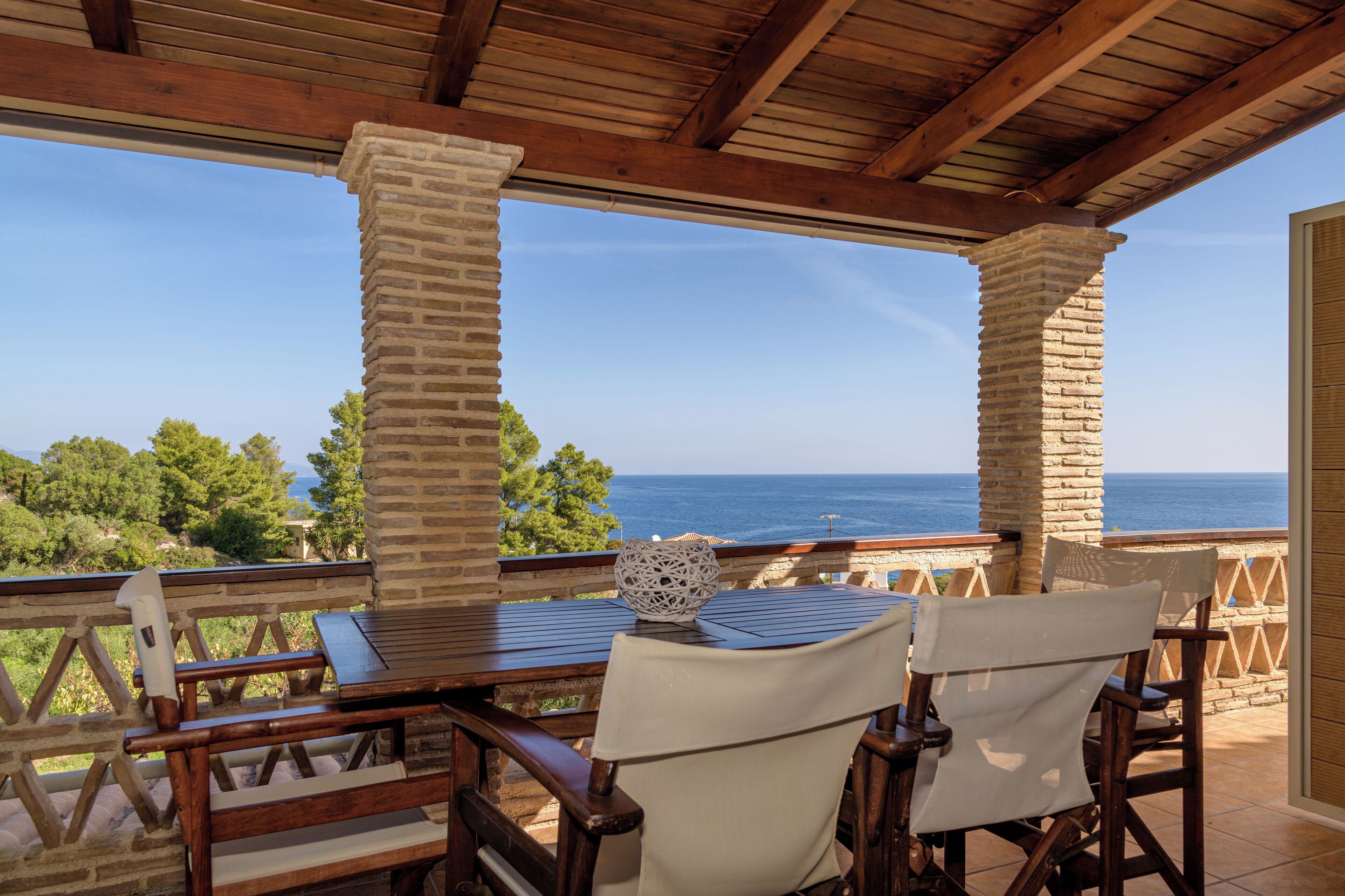 An outdoor patio with a wooden table and chairs, overlooking a scenic view of the sea and greenery.