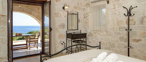 A bedroom with a bed, a nightstand, a dresser, a coat rack, and a view of the sea through an open door.