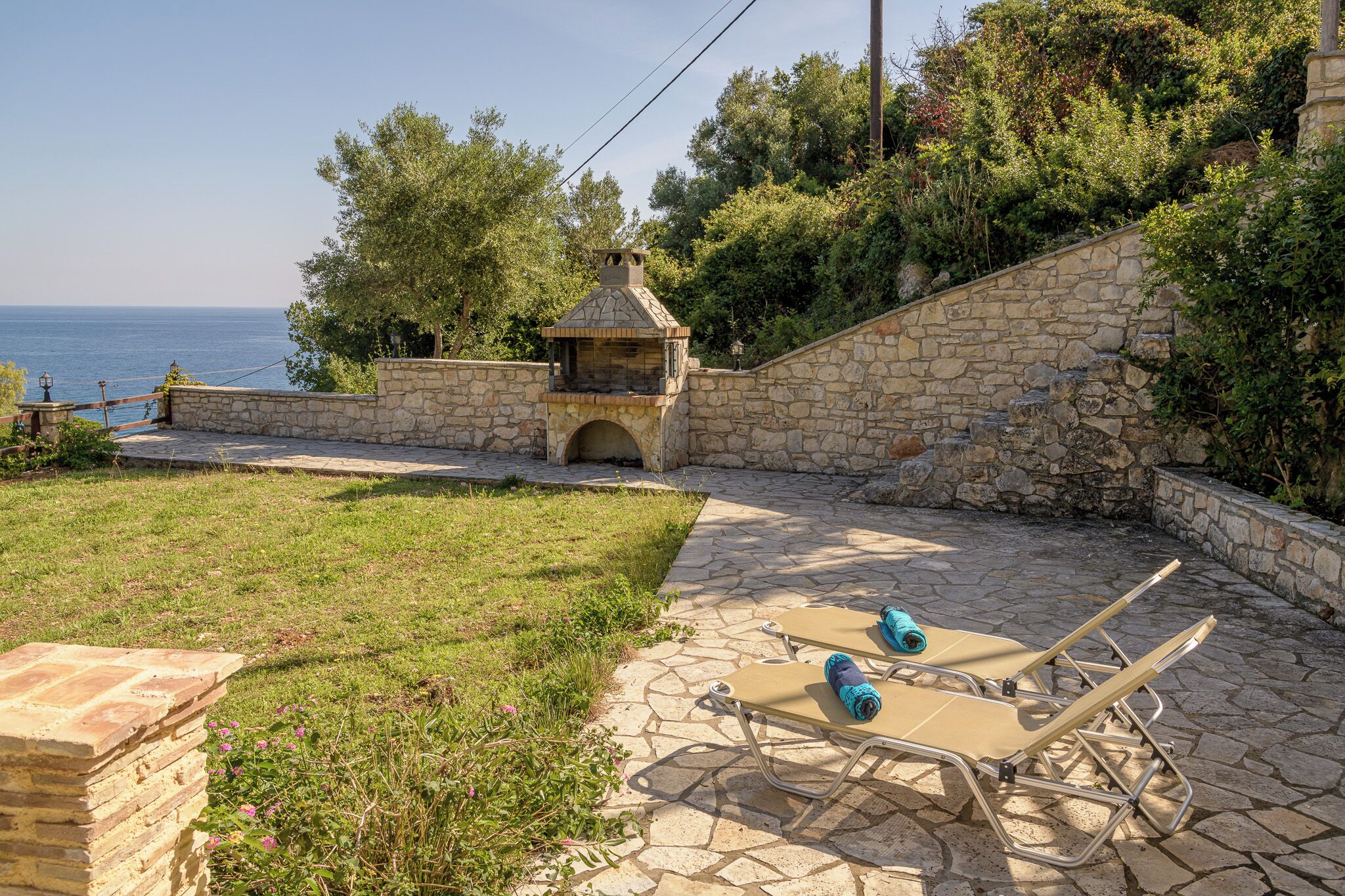 Un patio de piedra con sillones de descanso y una pequeña construcción de piedra, con vista a un cuerpo de agua.