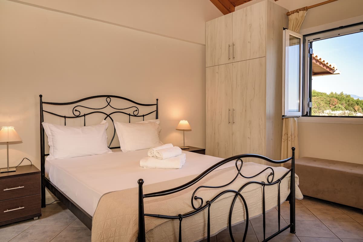 A neatly made bed with white linens, a bedside lamp, a wooden wardrobe, and a window with a view of greenery.