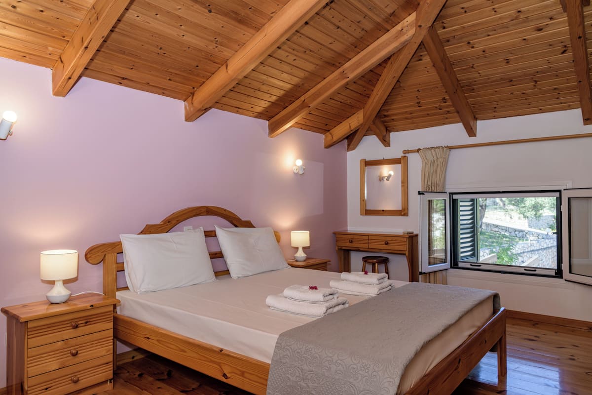 A bedroom with a wooden ceiling, a bed with white linens, a nightstand with a lamp, and a window with a view.