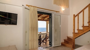 Habitación con escalera de madera, puerta corrediza de vidrio que da a un balcón con vistas al mar, una mesa con un florero y un televisor montado en la pared.