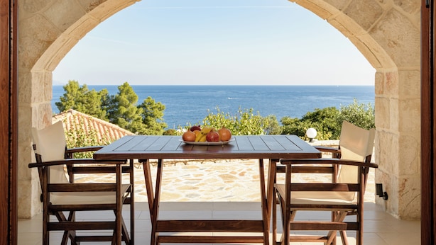 Un arco de piedra conduce a una mesa con frutero, con vistas a una playa y al mar.