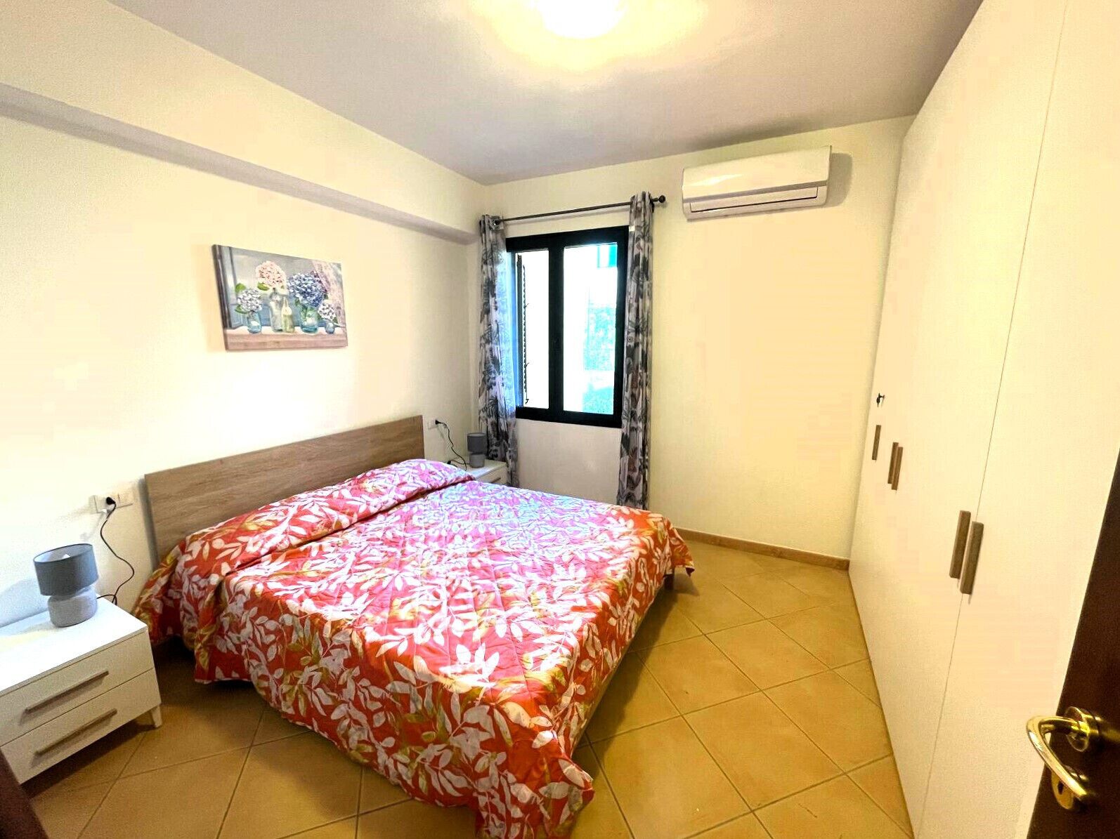 Ein Schlafzimmer mit Bett, Nachttisch, einem Fenster mit Vorhängen, einer Klimaanlage und einer Tür.