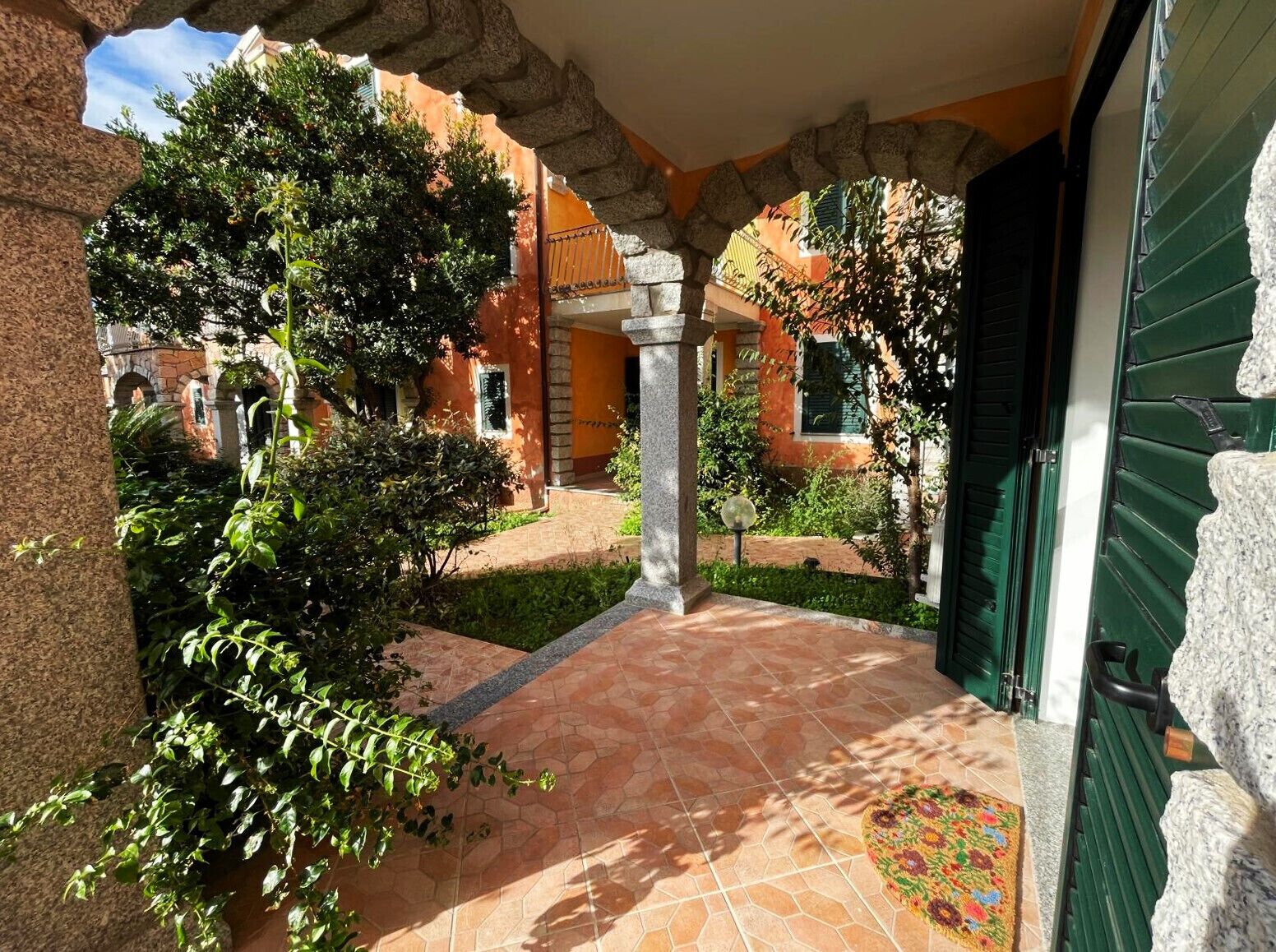 A covered outdoor area with a tiled floor, stone columns, and greenery.