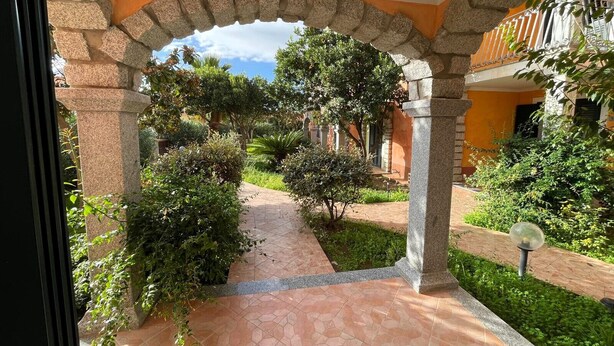 Un patio exterior con un arco de piedra, un sendero adoquinado y abundante vegetación.