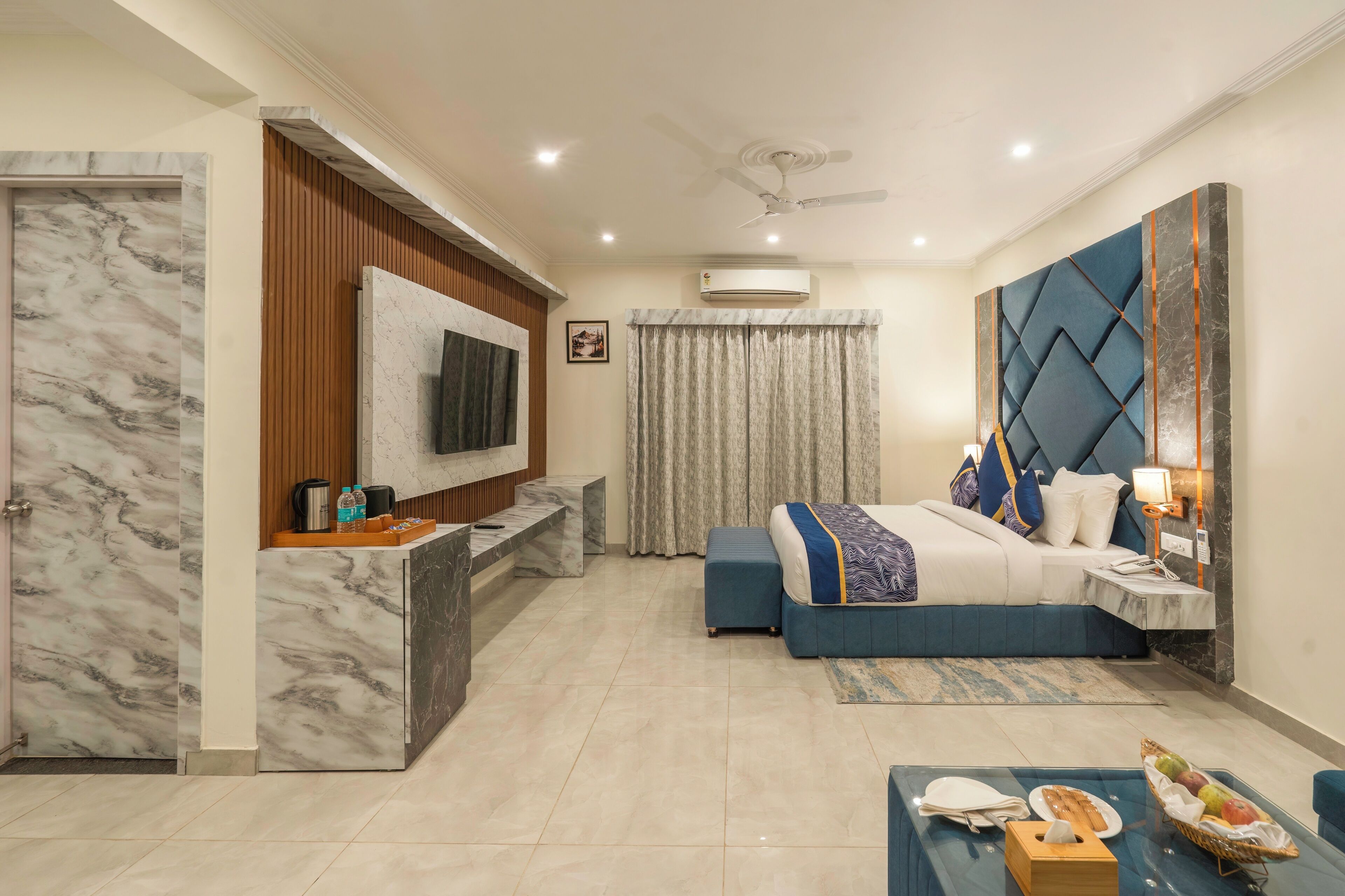 A hotel room with a marble wall, a bed with blue bedding, a TV mounted on the wall, a desk with a computer, and a window with curtains.