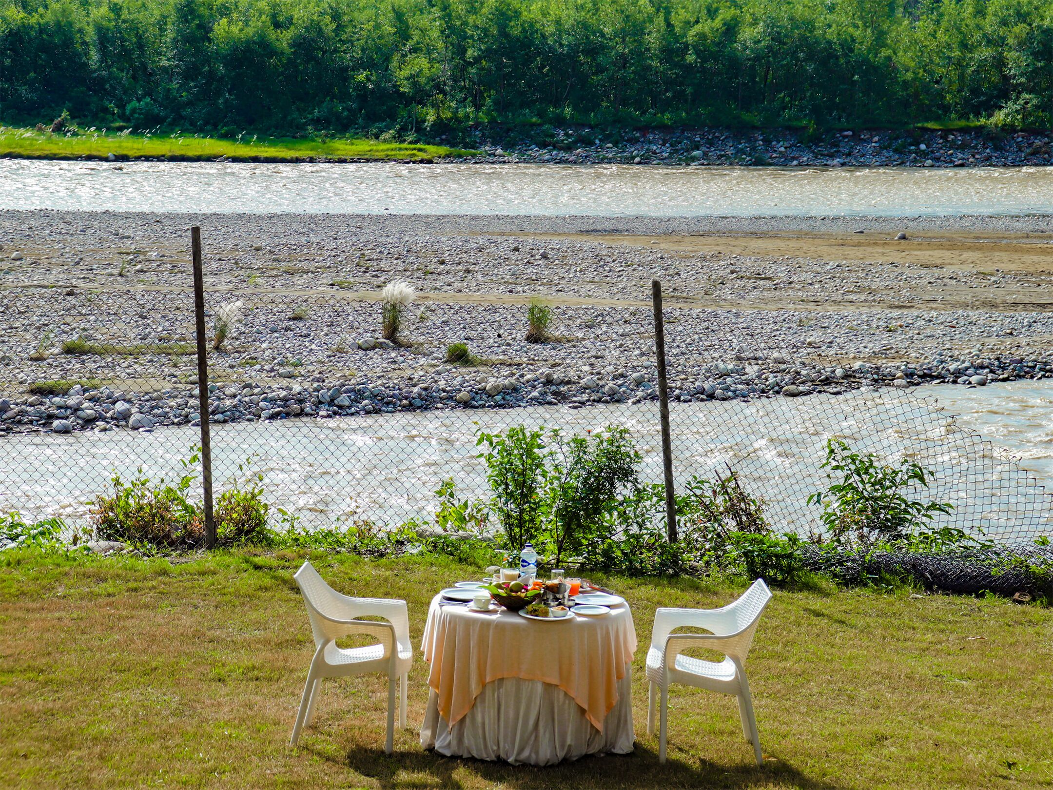 Una mesa de comedor con mantel y dos sillas dispuestas en una zona verde con vista a un río y un bosque al fondo.