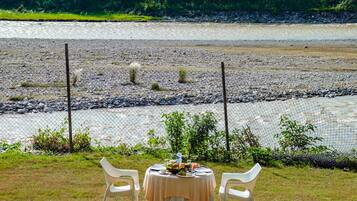 Una mesa de comedor con mantel y dos sillas dispuestas en una zona verde con vista a un río y un bosque al fondo.