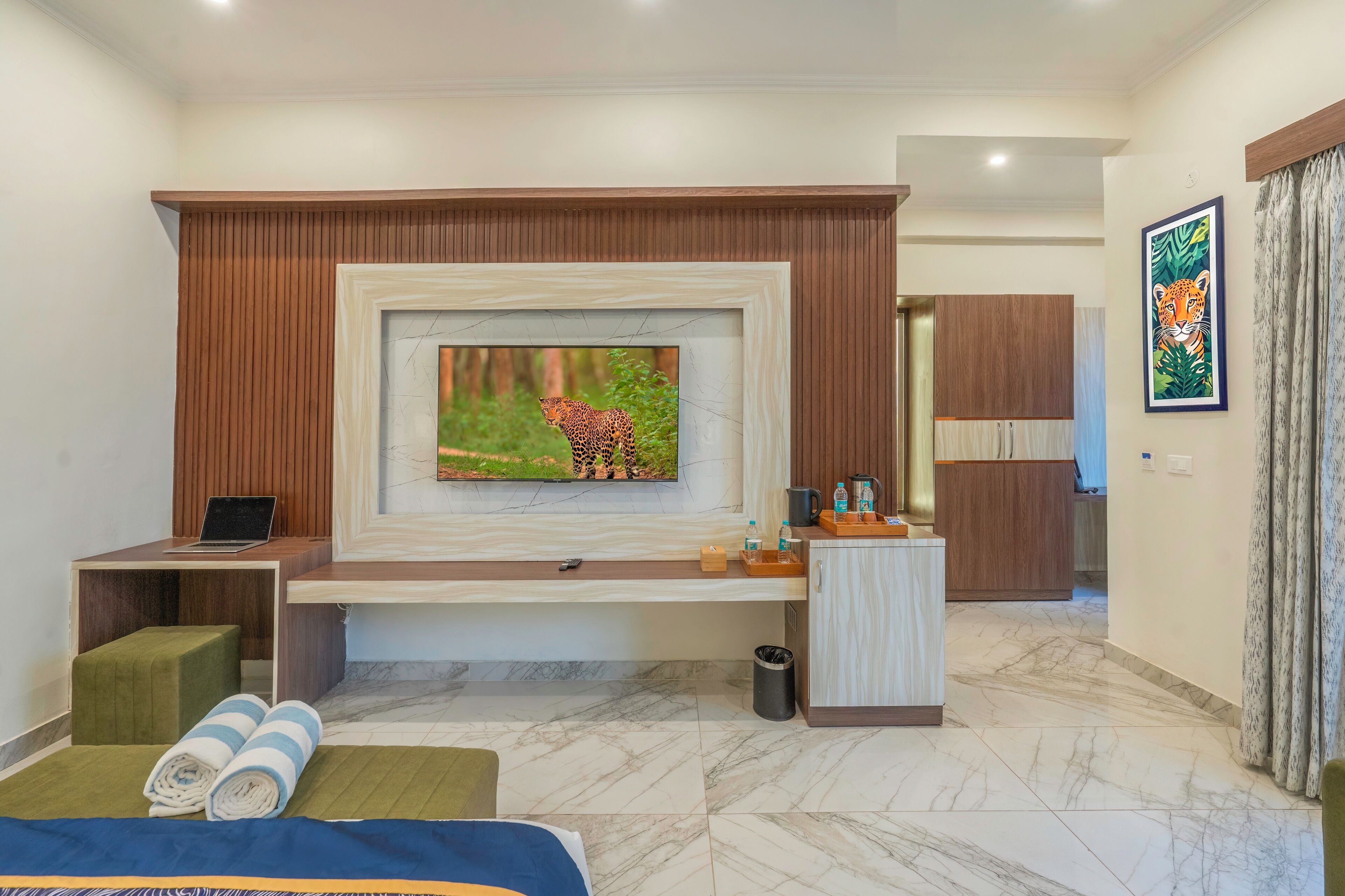 A modern hotel room with a large TV mounted on a wooden panel wall, a bed with green and blue bedding, a small desk with a laptop, a wooden cabinet, and a trash bin.