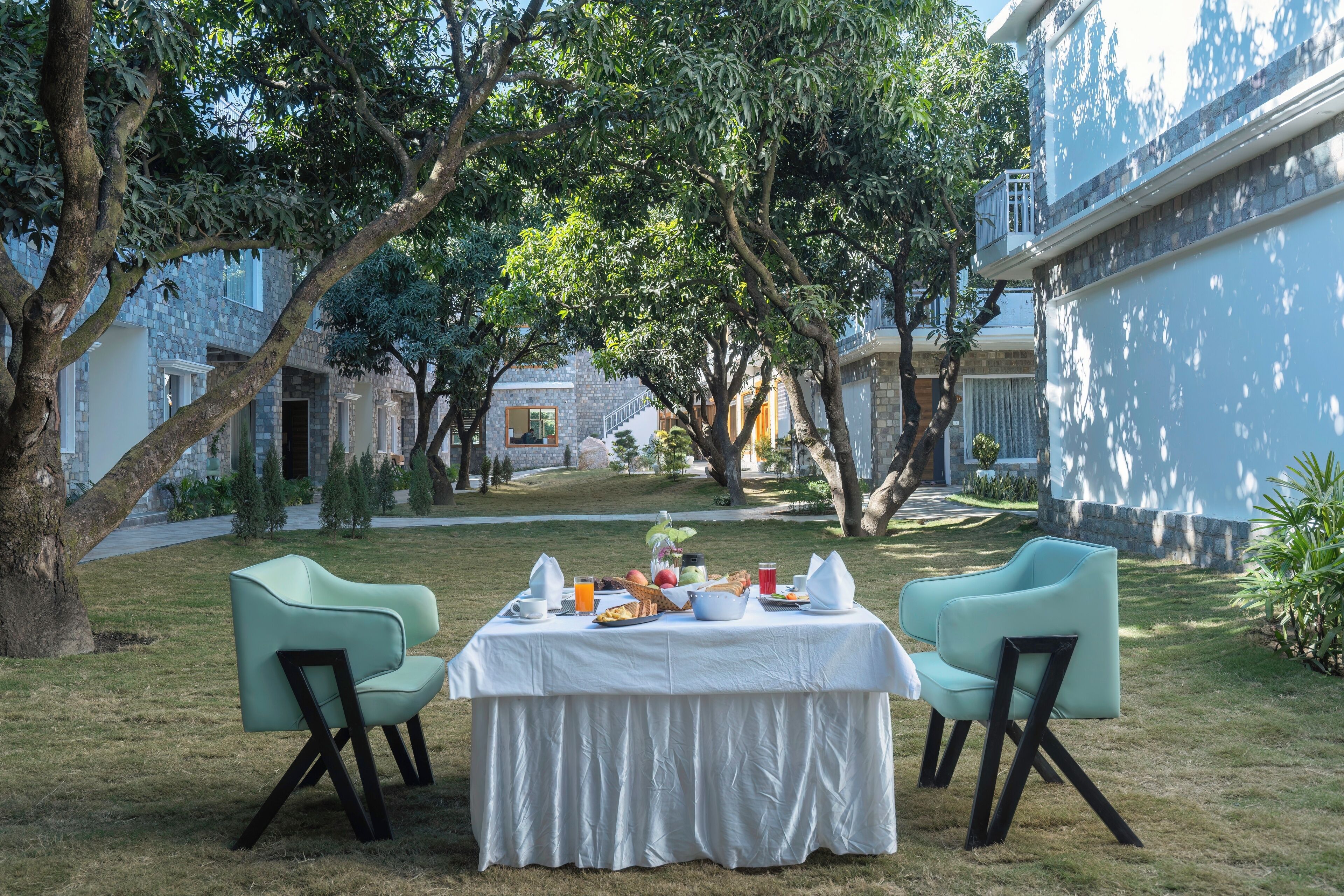 An outdoor dining area with a table set for breakfast, surrounded by trees and a building in the background.