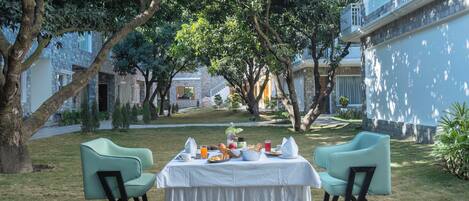 An outdoor dining area with a table set for breakfast, surrounded by trees and a building in the background.