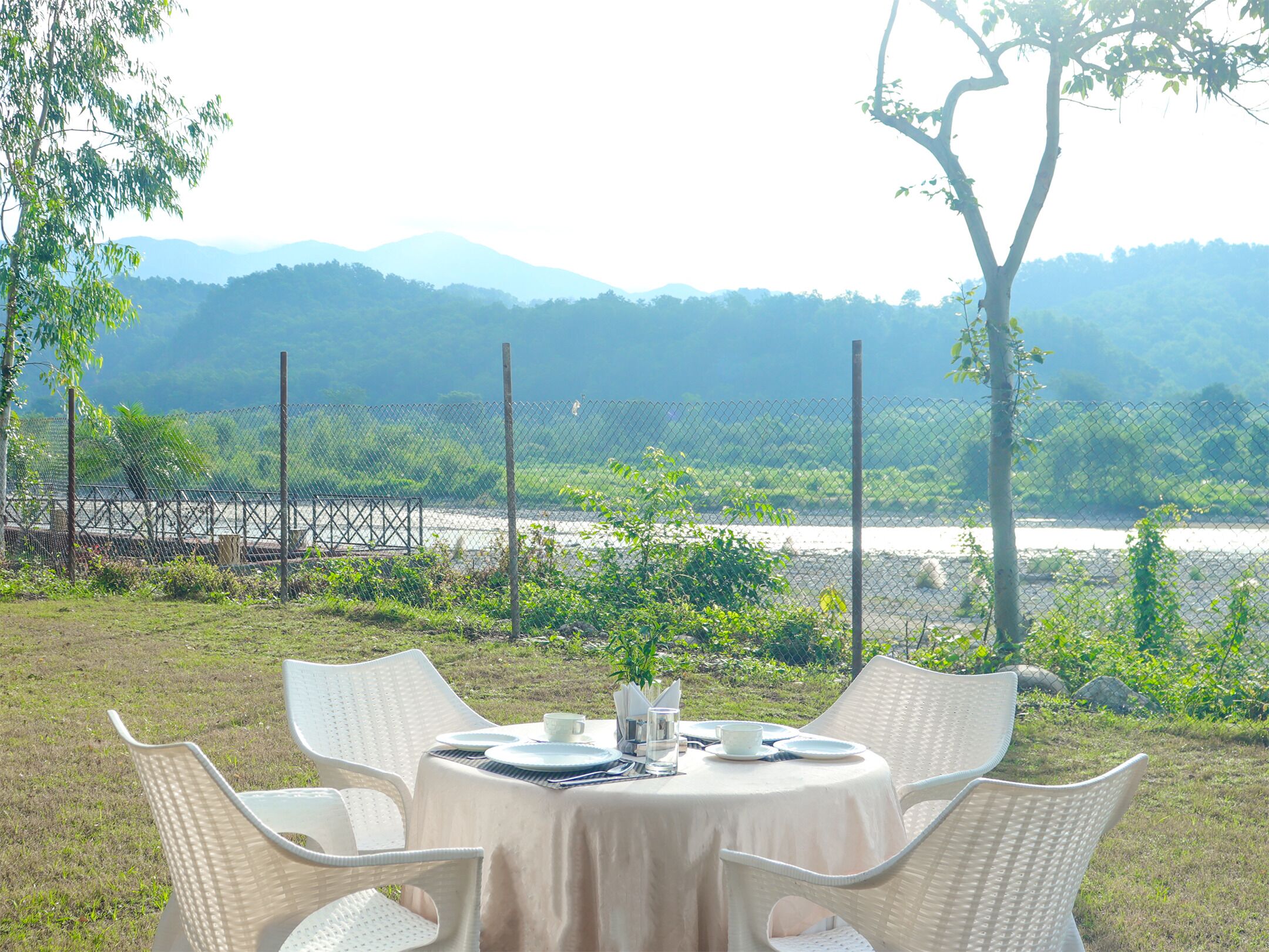 A round table with white wicker chairs set outdoors, overlooking a river and distant mountains.