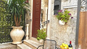 Un edificio in pietra con una porta rossa, una pianta in vaso, un tavolo con una bottiglia e dei bicchieri, e un cartello con un numero.