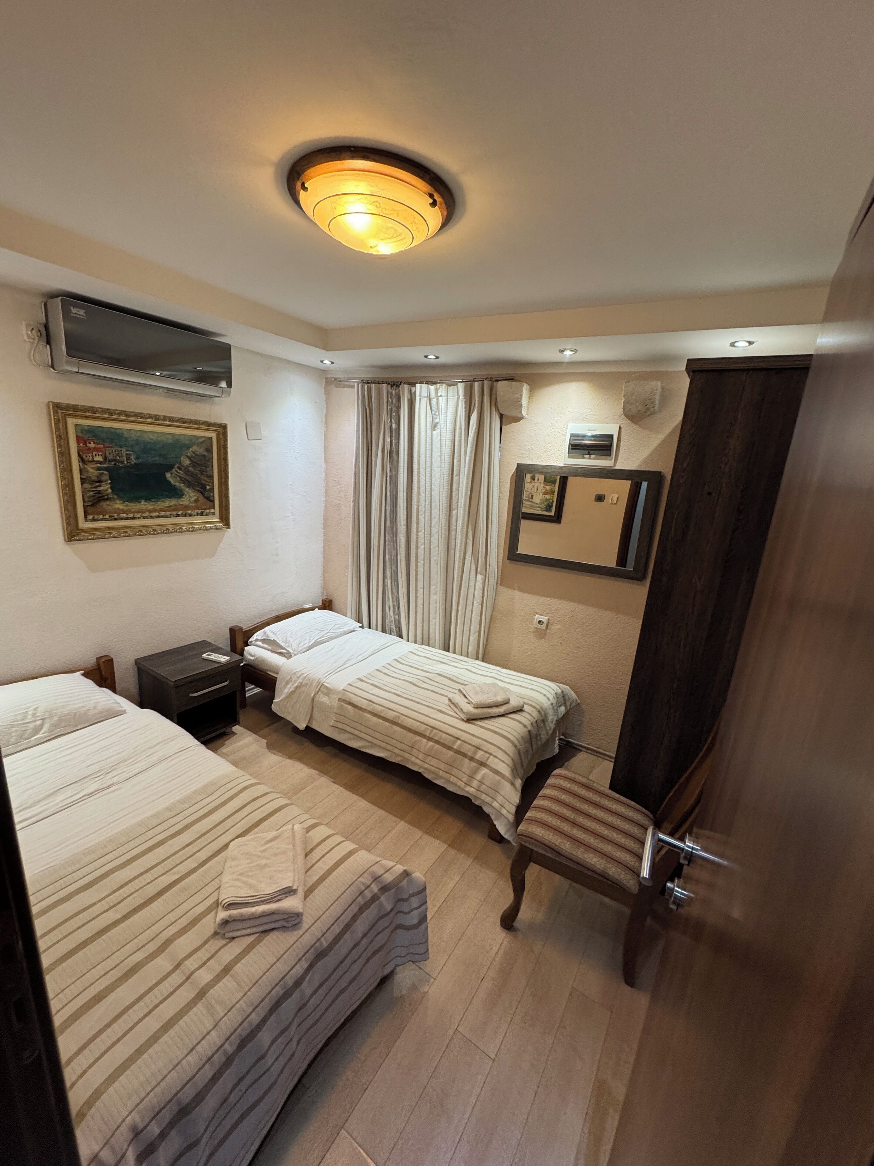 A hotel room with two single beds, a small bedside table, a chair, a wardrobe, a painting, and a ceiling light.