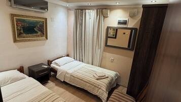 A hotel room with two single beds, a small bedside table, a chair, a wardrobe, a painting, and a ceiling light.