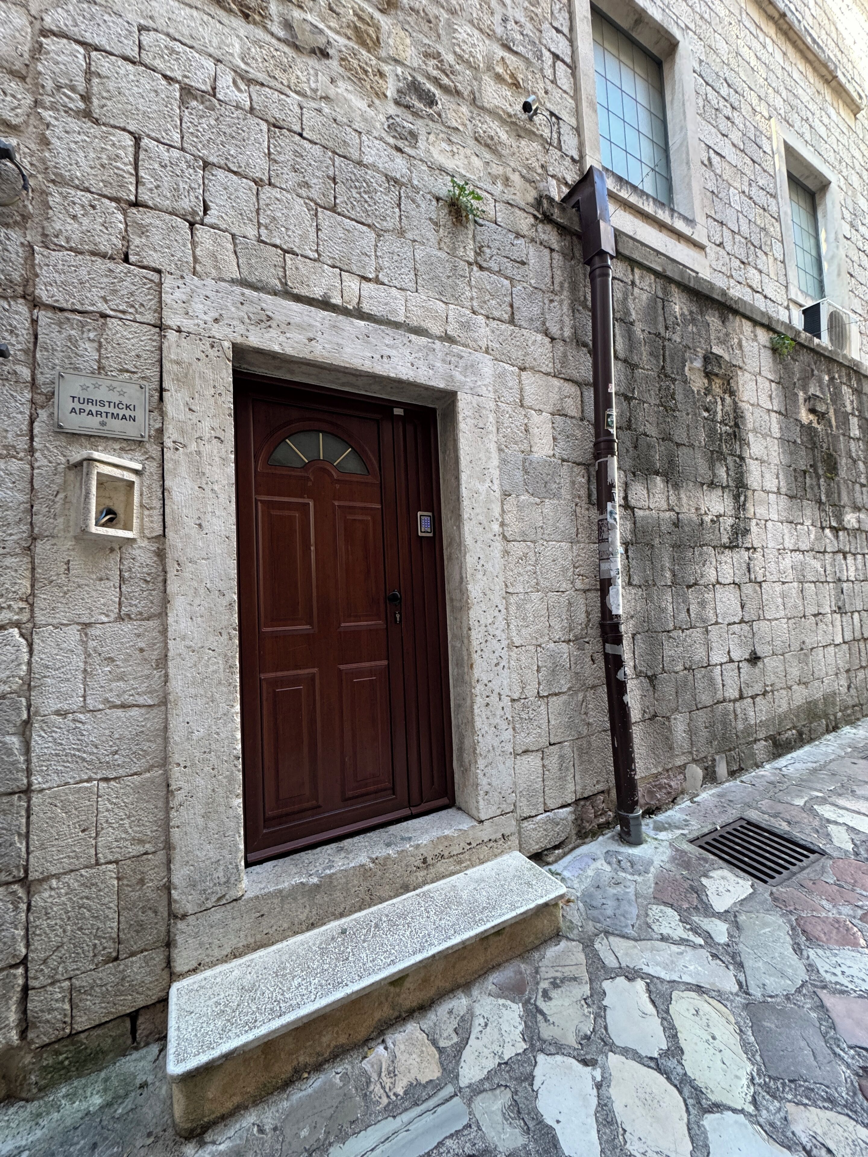 A stone building with a wooden door, a small sign above the door, and a window with a view of the sky.