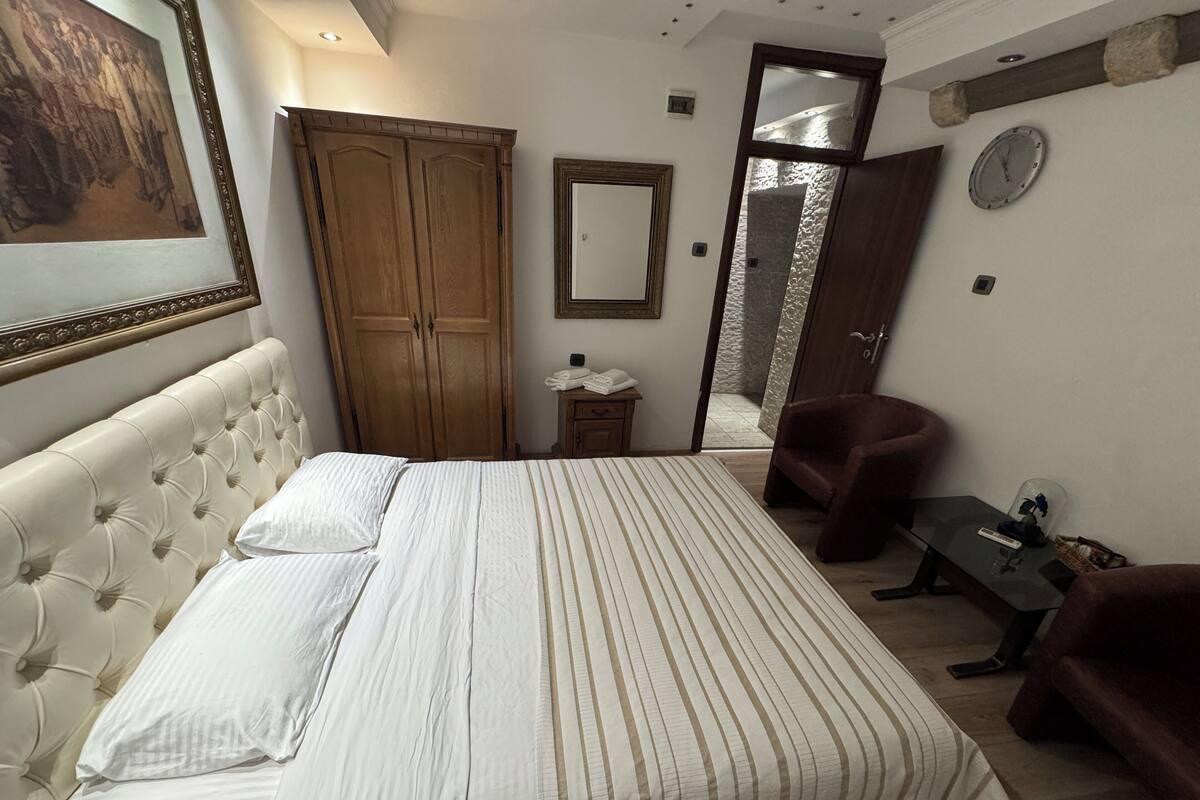 A bedroom with a large bed, a wooden wardrobe, a mirror, a chair, and a small table.