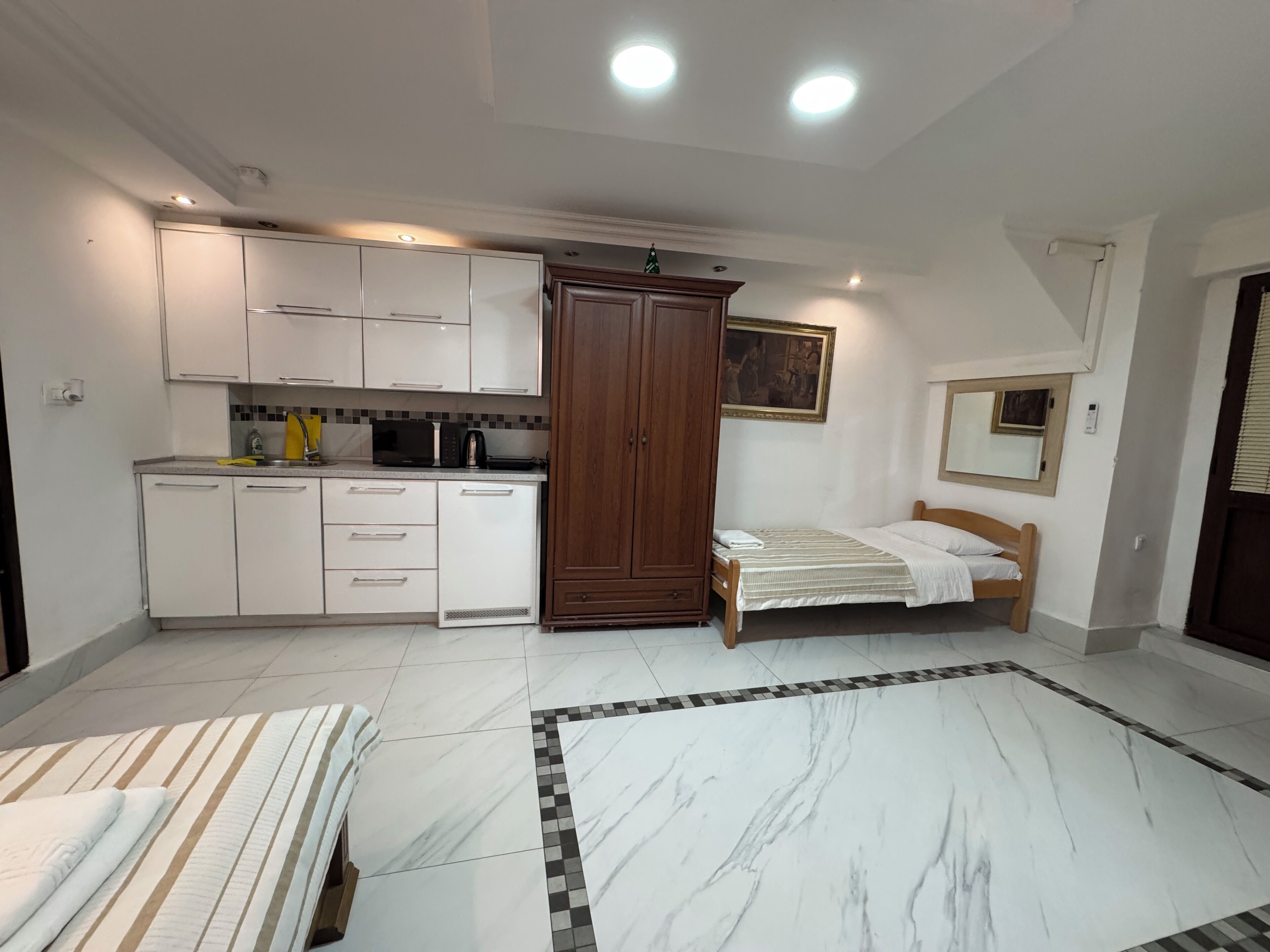 A bedroom with a bed, a small bedside table, a wooden wardrobe, and a kitchen area with cabinets and appliances.