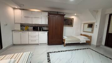 A bedroom with a bed, a small bedside table, a wooden wardrobe, and a kitchen area with cabinets and appliances.