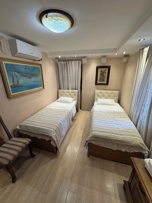 A hotel room with two single beds, a bench, a painting, and an air conditioning unit. - Vita Mundo (Kotor)