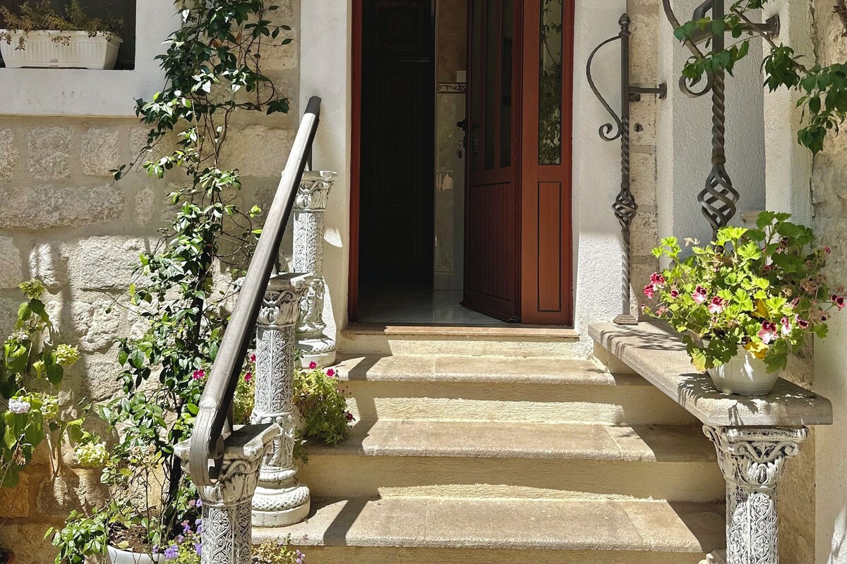 Una escalera de piedra que lleva a una puerta de madera con el número '2' sobre ella, flanqueada por macetas con plantas y columnas decorativas.
