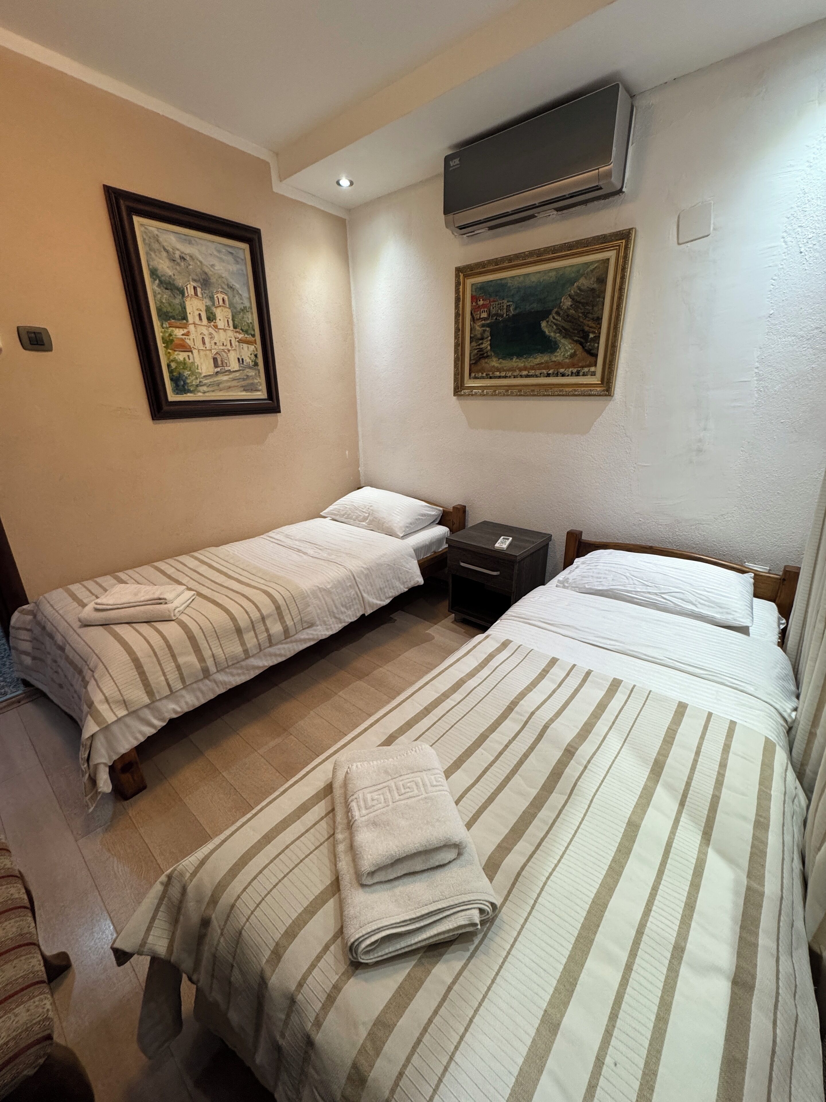 A room with two single beds, a nightstand, a small table, and a wall-mounted air conditioner.