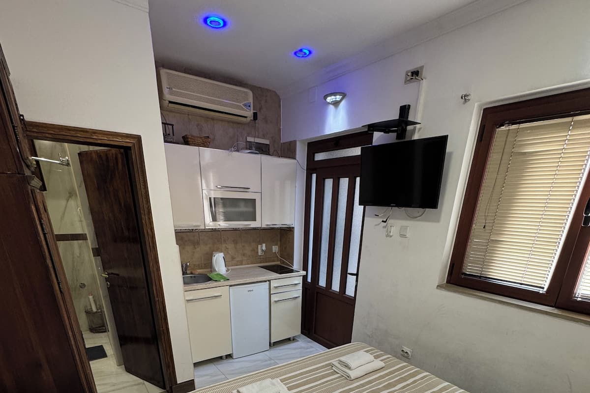 A compact bedroom with a bed, bedside table, and a small bedside lamp. A kitchen area is visible in the background with a microwave and a sink.
