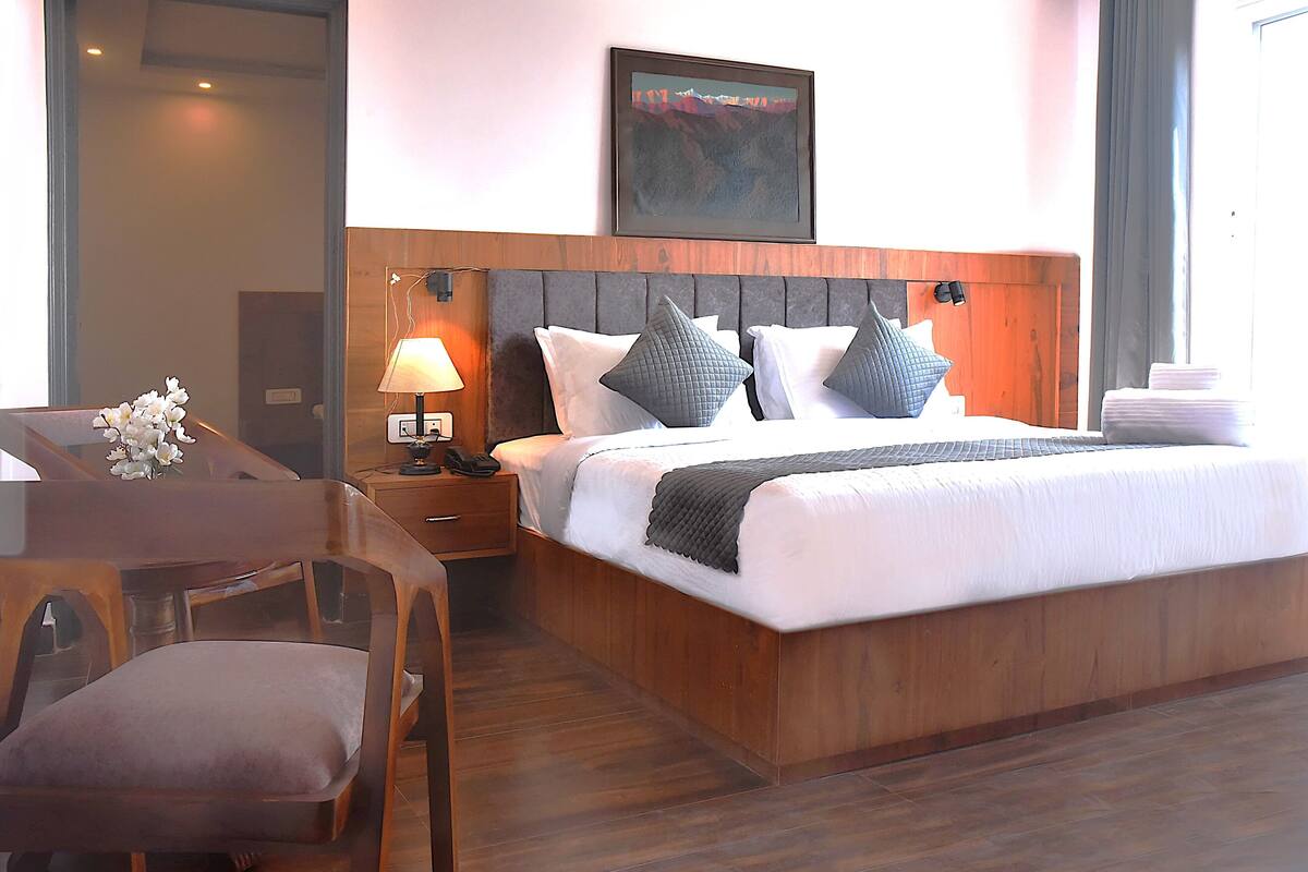Habitación de hotel con cabecera de madera, cama grande con ropa blanca y almohadas grises, mesita de noche con lámpara y un cuadro en la pared.