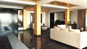 Een ruime woonkamer met een grote bank, houten balken, een flatscreen televisie en een eethoek met tafel en stoelen.
