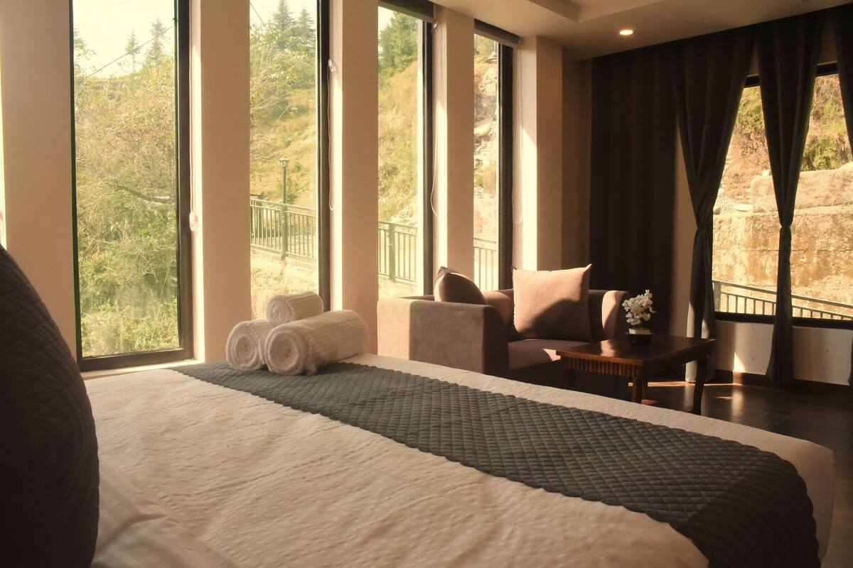 Habitación de hotel con una cama grande, una zona de estar con una mesa y vistas al exterior a través de amplios ventanales.